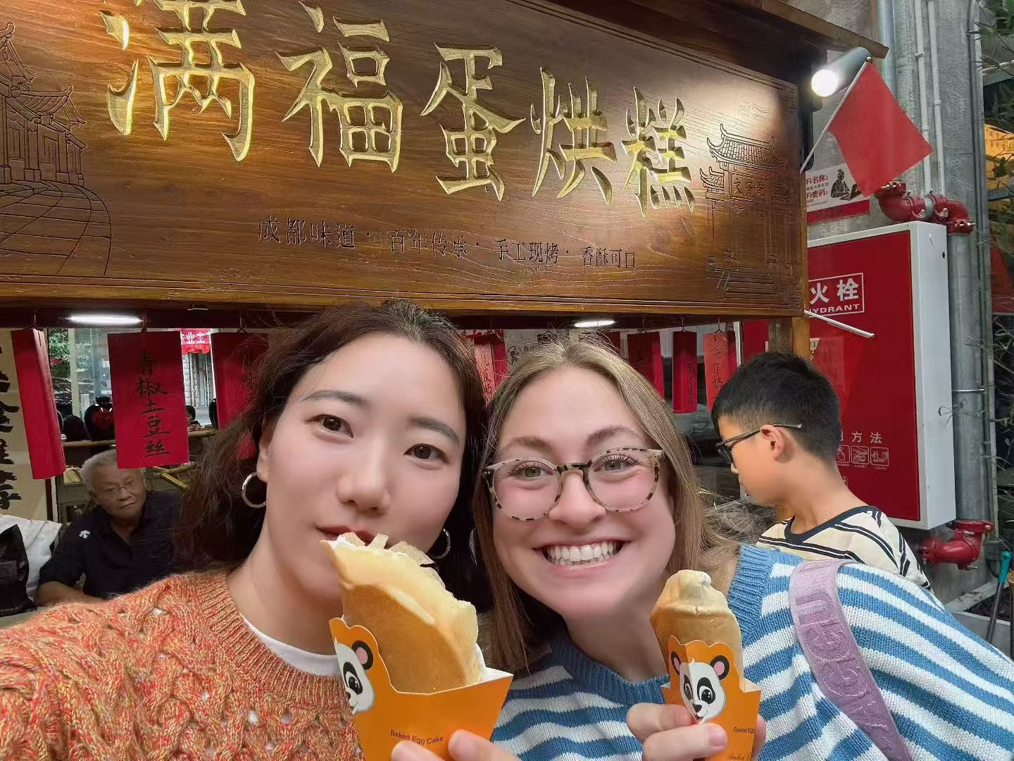 Two women holding baked egg cakes outside a street-food stall.