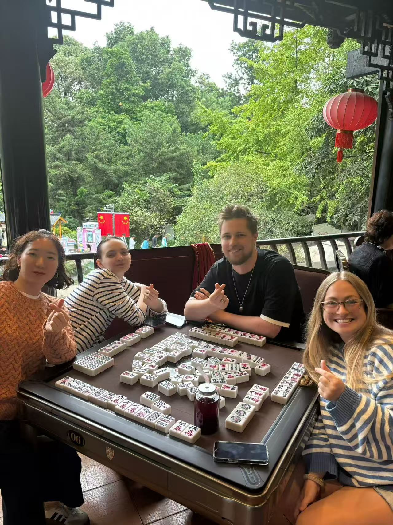 Four people smiling around a mahjong table beside an open window overlooking trees.