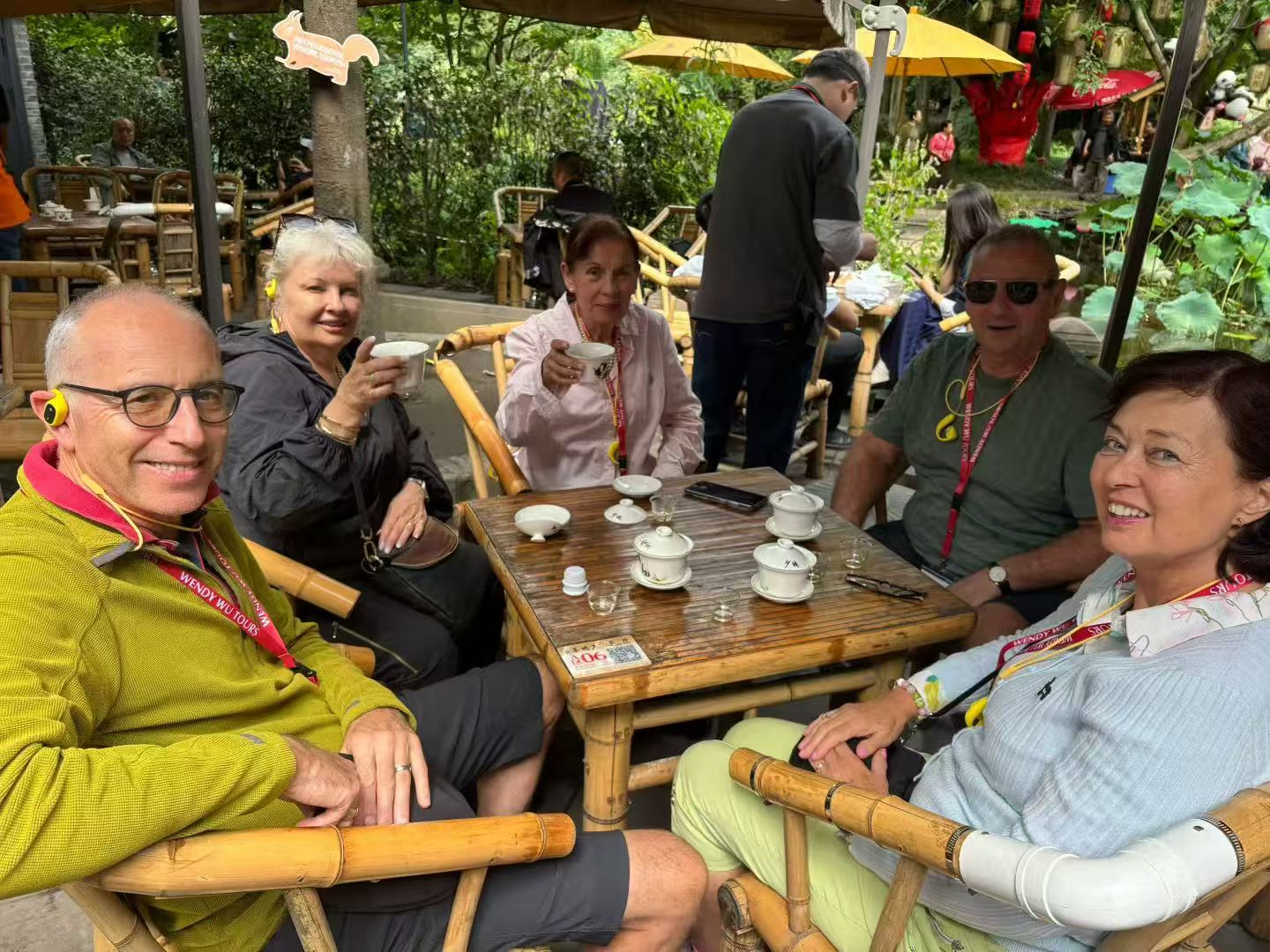 Five guests seated around a tea table on bamboo chairs in a leafy outdoor teahouse.