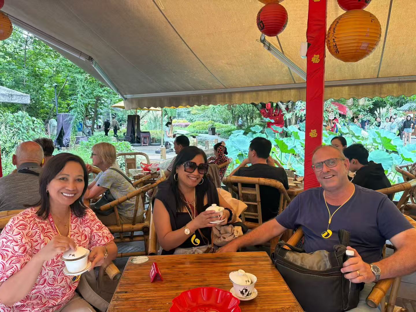 Three smiling guests with lidded teacups at a wooden table beneath red and yellow lanterns.
