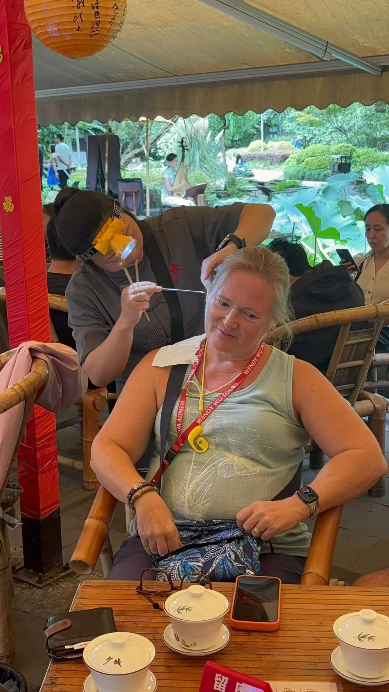 A woman seated in a bamboo chair while a practitioner uses a headlamp and slender tools for ear cleaning.