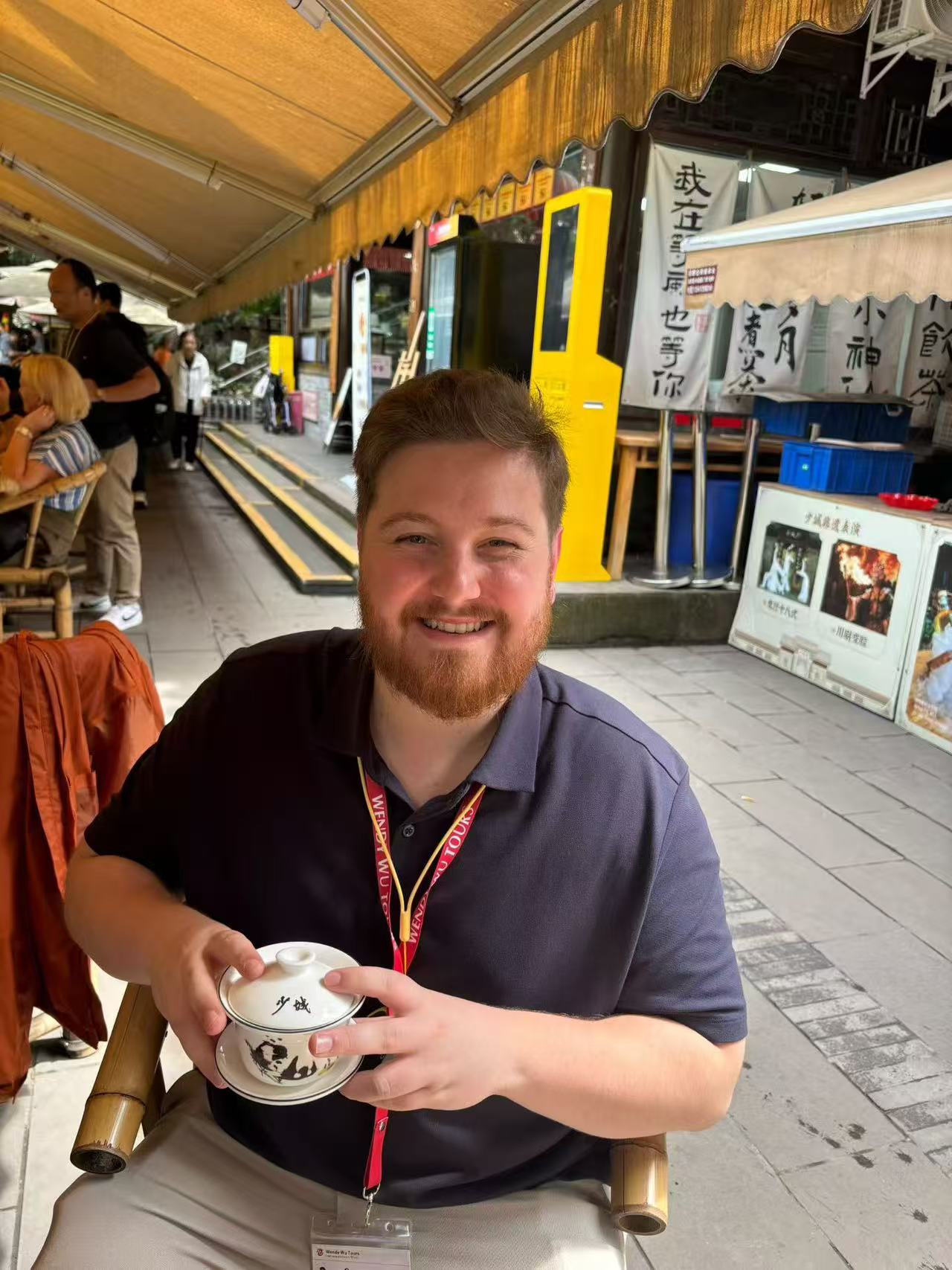 A smiling guest holding a lidded Chinese teacup while seated at a teahouse.
