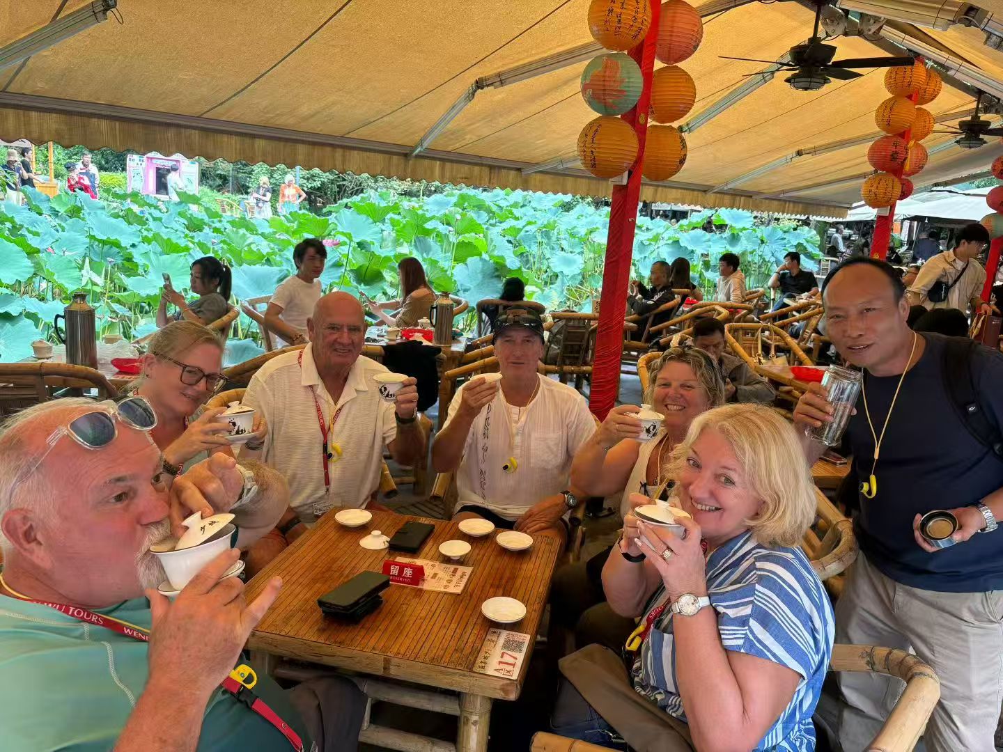 A group raising teacups around a bamboo table beneath lanterns, with lotus leaves behind them.