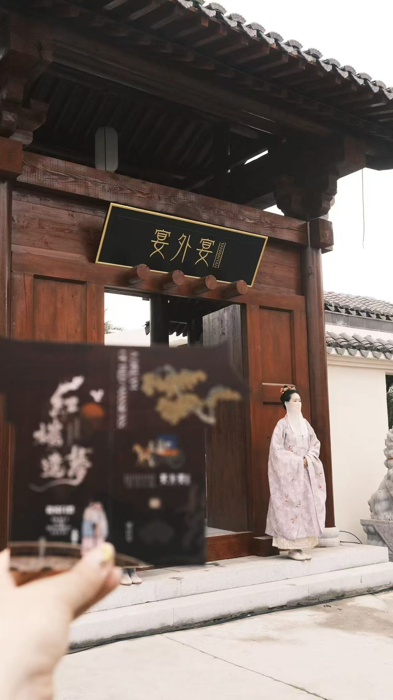 A hand holding a booklet in front of a wooden entrance, with a person in pale traditional dress beside the doorway.