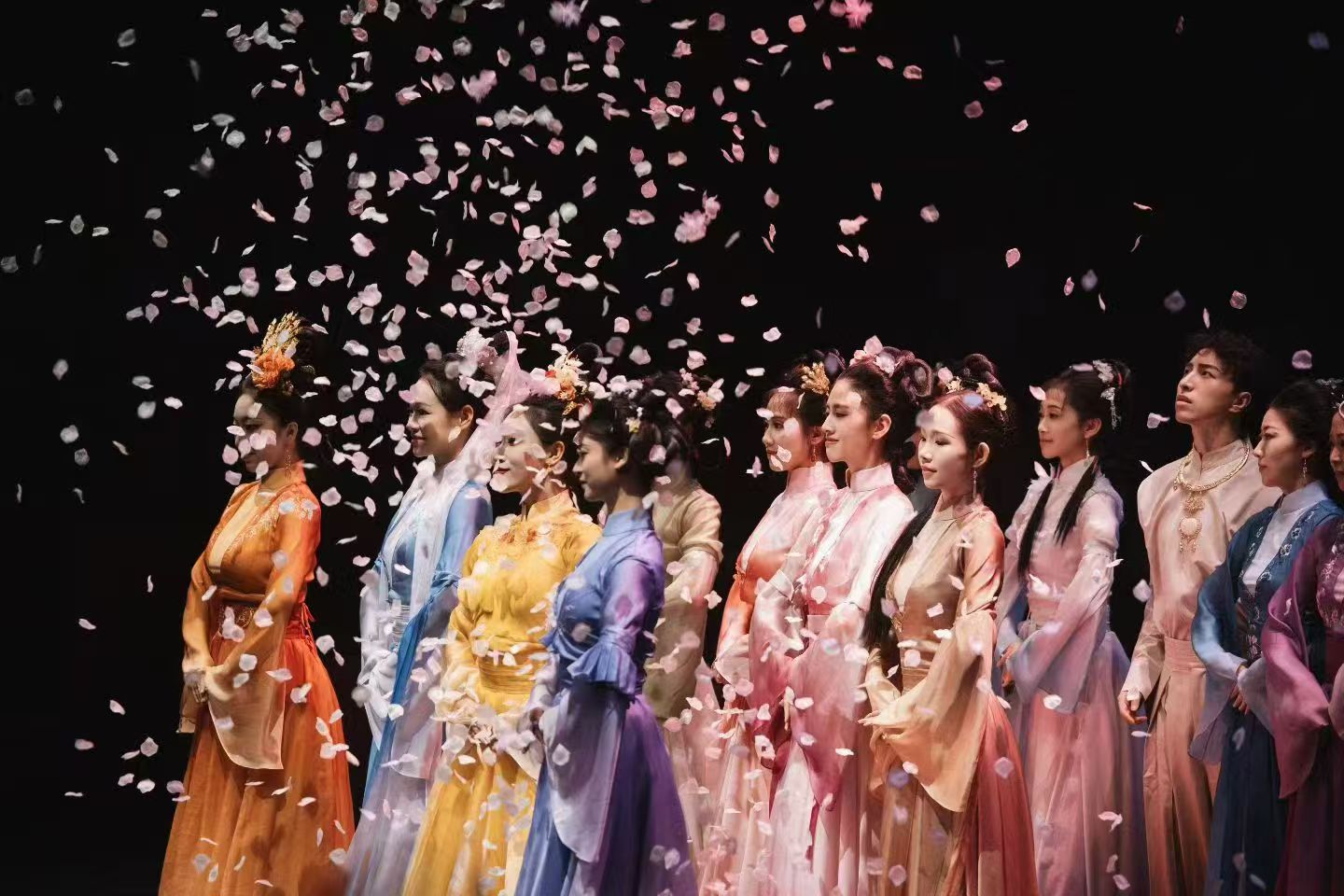 A row of performers in pastel traditional costumes standing beneath falling pink and white petals.