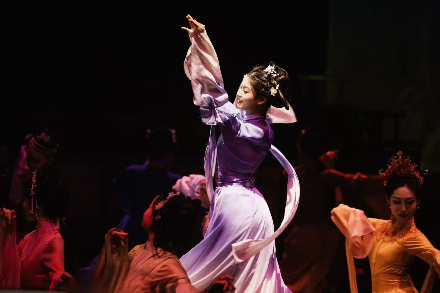 A dancer in a flowing purple costume raising one arm, with other performers behind her.