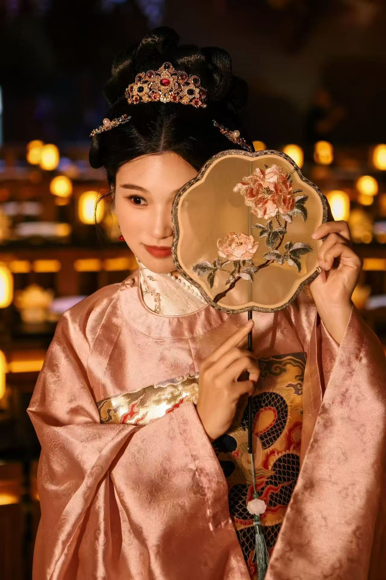 A woman in pink traditional dress holding a floral embroidered fan beside her face, with warm lanterns behind her.