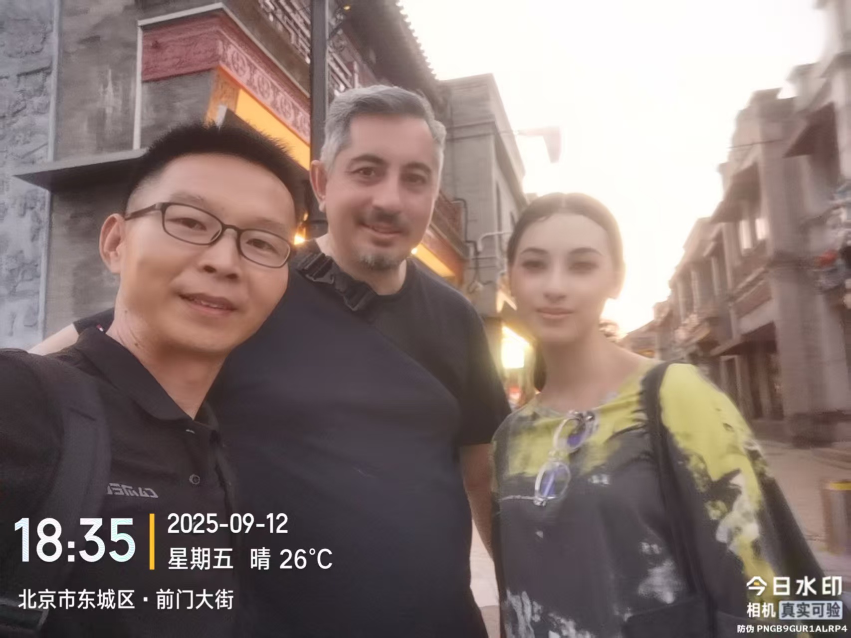 Three people taking a selfie on a street lined with grey brick buildings in the evening light.