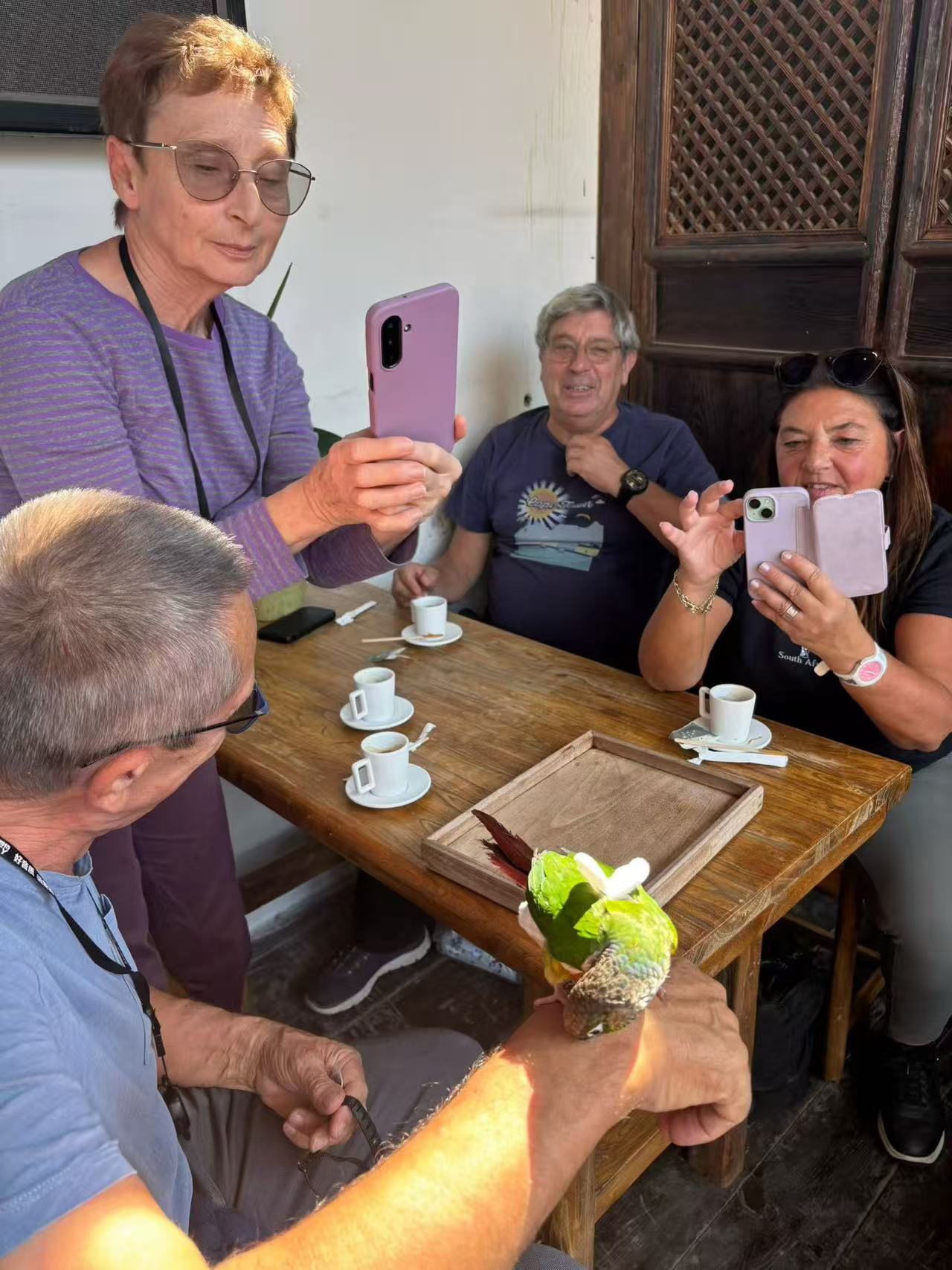 Guests taking photos of a small green parrot perched on an arm beside a wooden tea table.