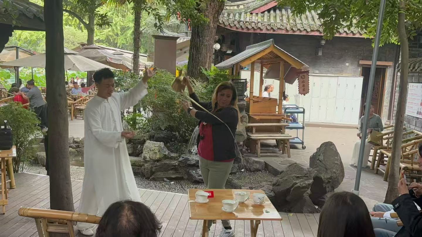 A person in a long white robe demonstrating tea service beside a guest in a shaded courtyard teahouse.