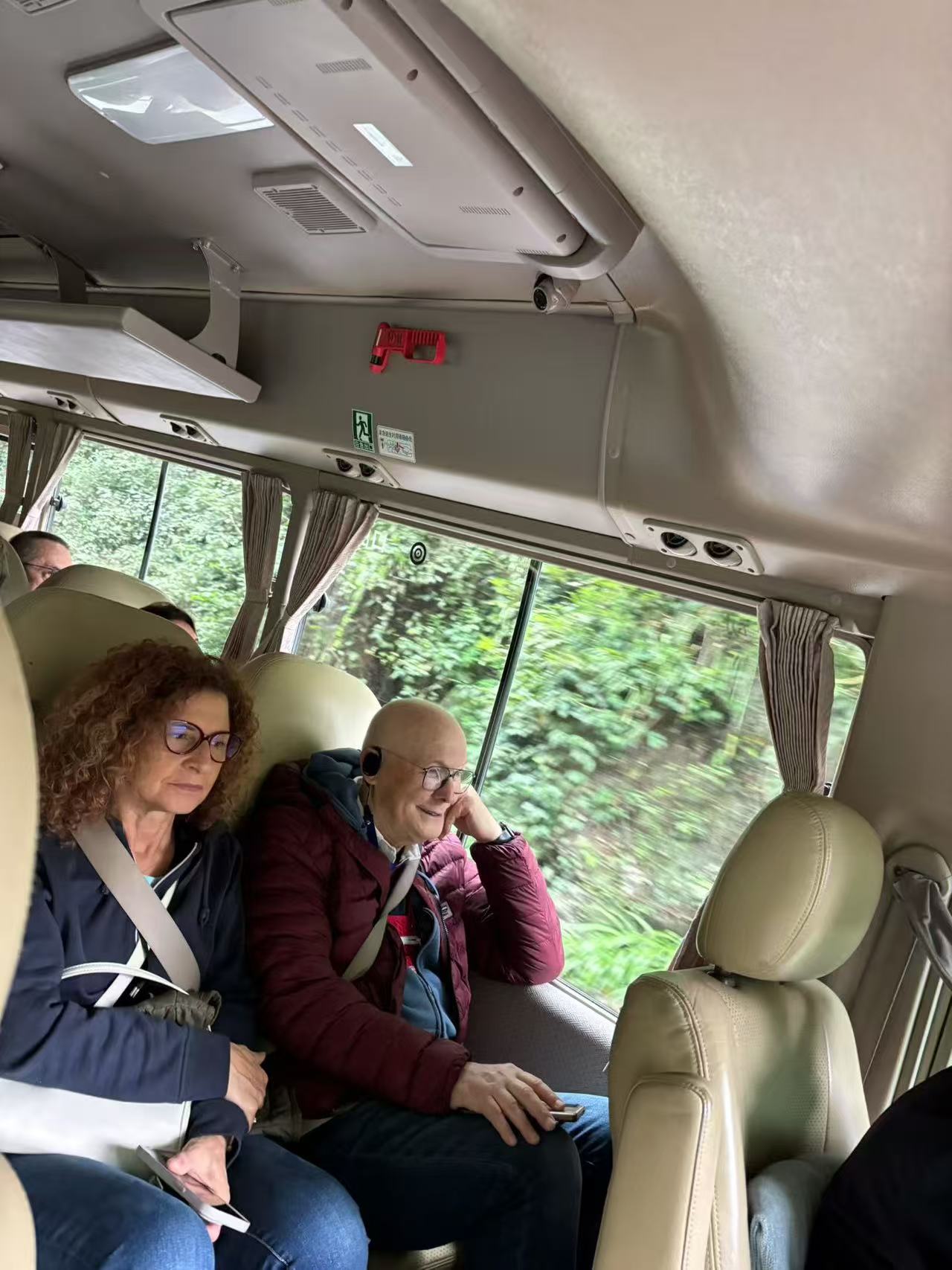 Two guests seated beside a minibus window overlooking green trees.
