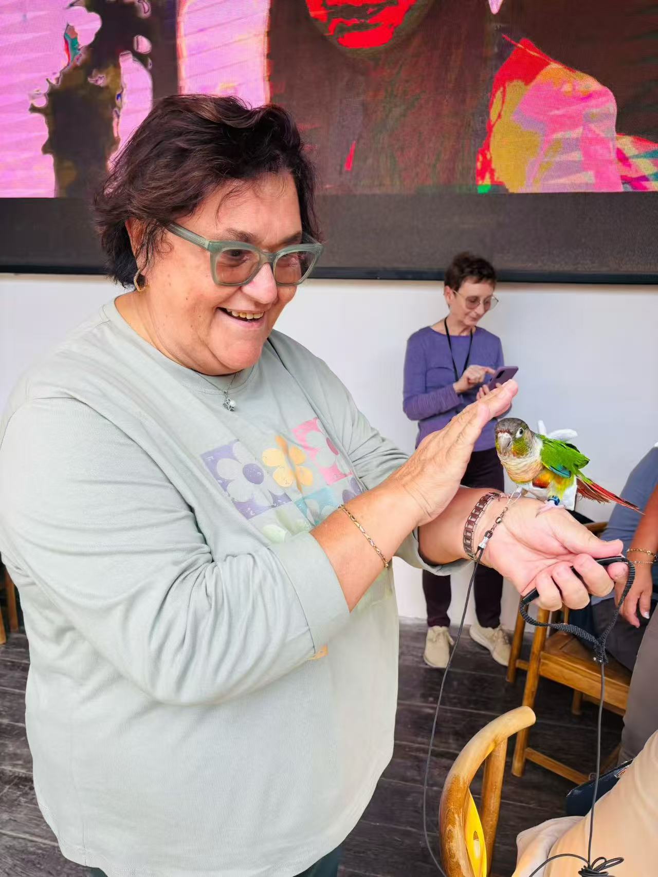 A smiling guest holding a small green parrot on one hand and gently reaching toward it with the other.