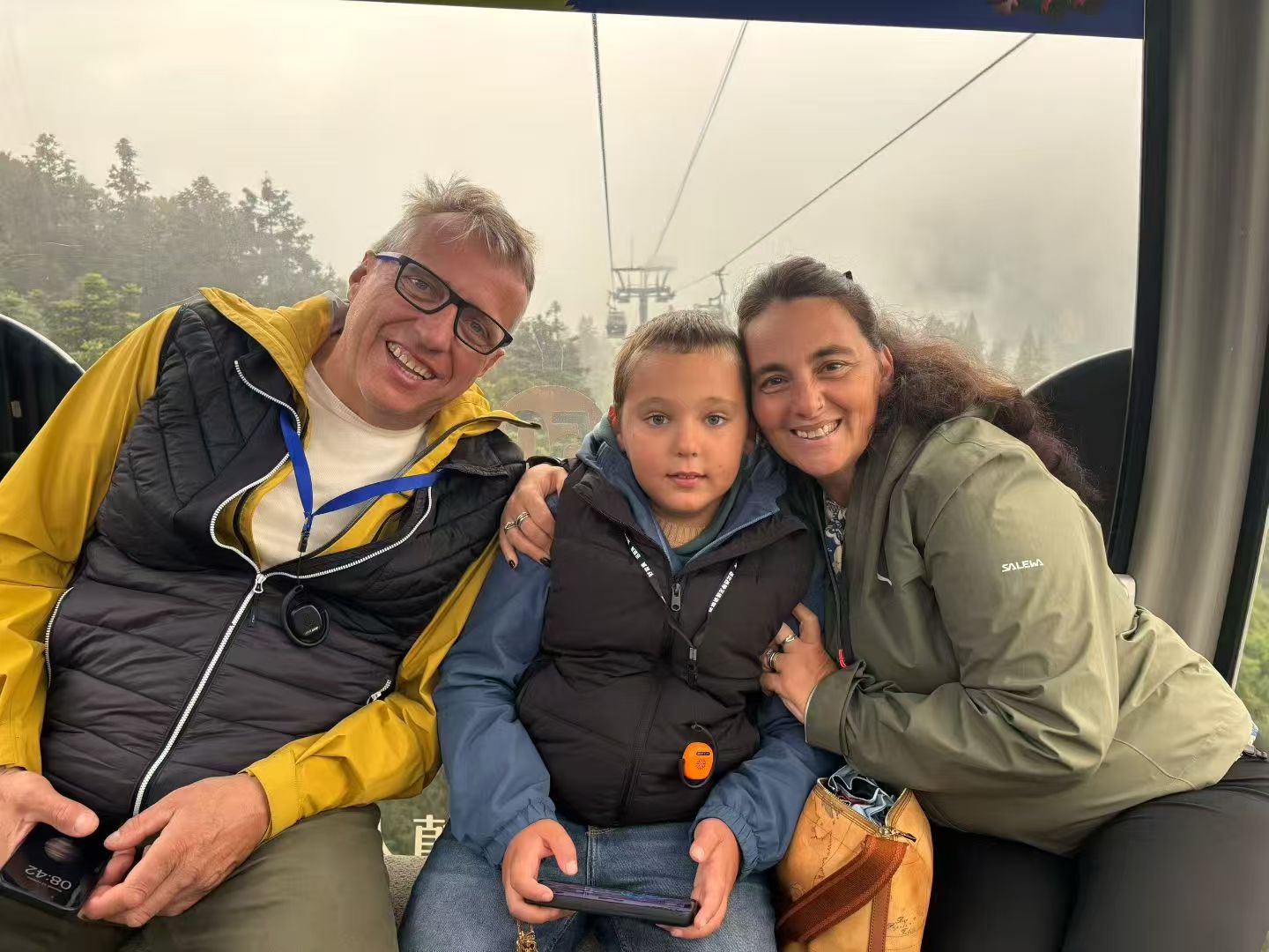 Two adults and a child smiling together inside a cable car, with misty trees visible through the window.
