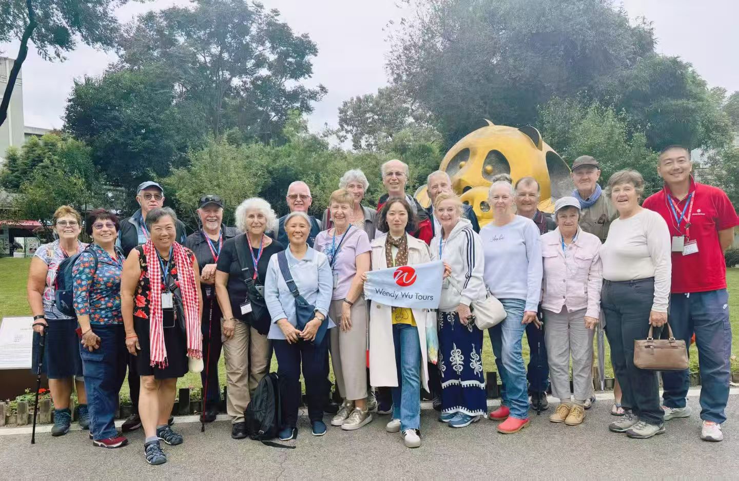 A large tour group posing outdoors in front of a golden panda sculpture and leafy trees.