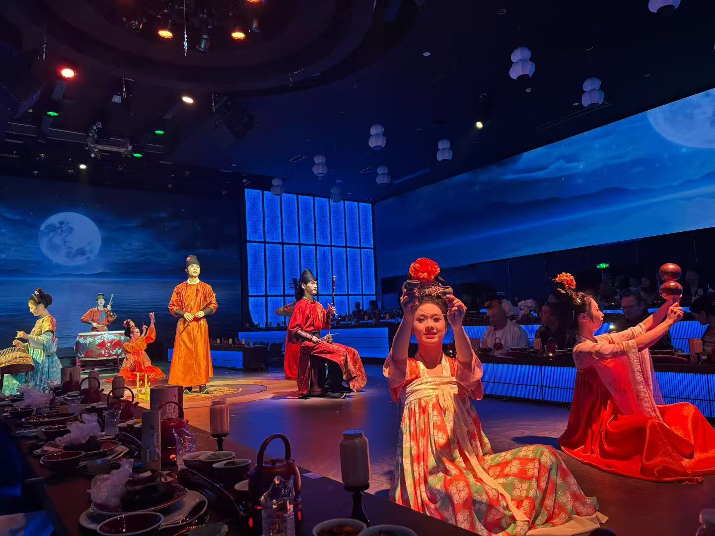 Performers in orange, red and green traditional costumes on a circular stage under blue and purple lights.